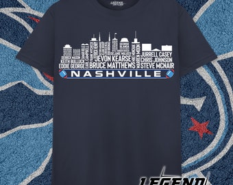 Może przedstawiać: Koszulka w kolorze granatowym z białą sylwetką panoramy Nashville. Panorama składa się z nazwisk znanych graczy futbolu amerykańskiego, w tym Eddiego George'a, Steve'a McNaira i Jevon Kearse'a. Pod panoramą znajduje się napis "NASHVILLE".