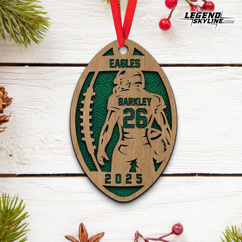 May include: Wooden football-shaped ornament with a green interior and a red ribbon. The ornament features the word "EAGLES" at the top, the name "BARKLEY" and the number "26" in the centre, and the year "2025" at the bottom. A sports-themed item.