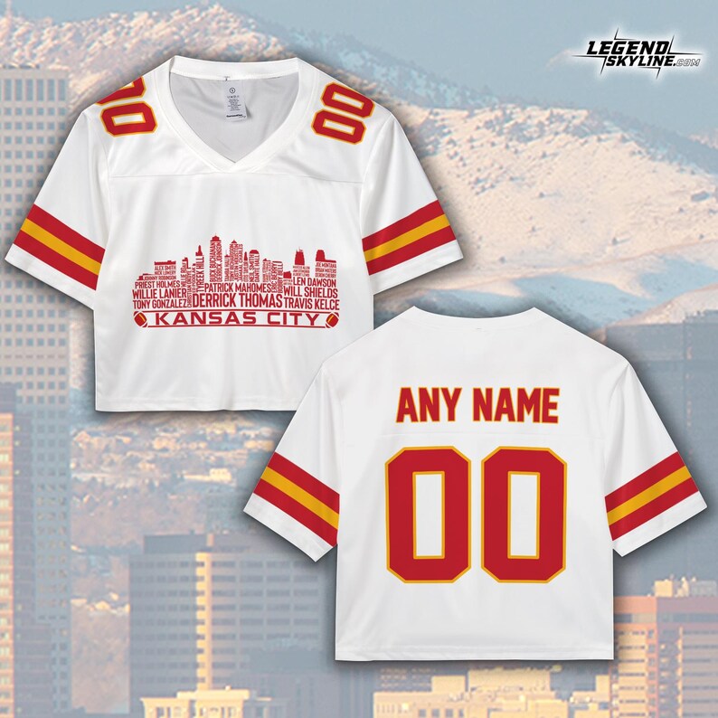 Puede incluir: Camiseta corta blanca con detalles rojos y amarillos. Presenta "KANSAS CITY" y un gr&aacute;fico de horizonte con nombres de jugadores. La parte trasera muestra "ANY NAME" y el n&uacute;mero "00".
