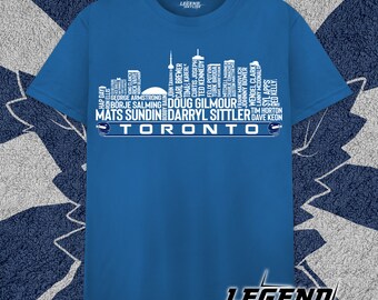 Puede incluir: Una camiseta azul con una silueta blanca del horizonte de Toronto. El horizonte está formado por los nombres de los famosos jugadores de hockey de los Toronto Maple Leafs. El texto "TORONTO" está escrito debajo del horizonte.
