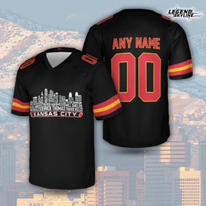 Puede incluir: Camiseta de f&uacute;tbol negra con detalles rojos y amarillos. La parte delantera presenta un gr&aacute;fico de horizonte blanco con nombres de jugadores y el texto "KANSAS CITY". La parte trasera muestra "ANY NAME" encima del n&uacute;mero "00".