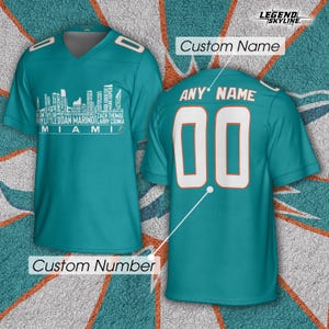 Puede incluir: Camiseta de fútbol turquesa con detalles blancos. La parte delantera presenta un gráfico de horizonte con nombres de jugadores y "MIAMI". La parte trasera muestra "ANY NAME" sobre el número "00". La camiseta es personalizable.