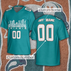 May include: A teal V-neck jersey featuring a white Miami skyline graphic with player names, the text MIAMI, and the number 00 on the front. The back displays the text ANY NAME and the number 00, with labels indicating Custom Name and Custom Number.