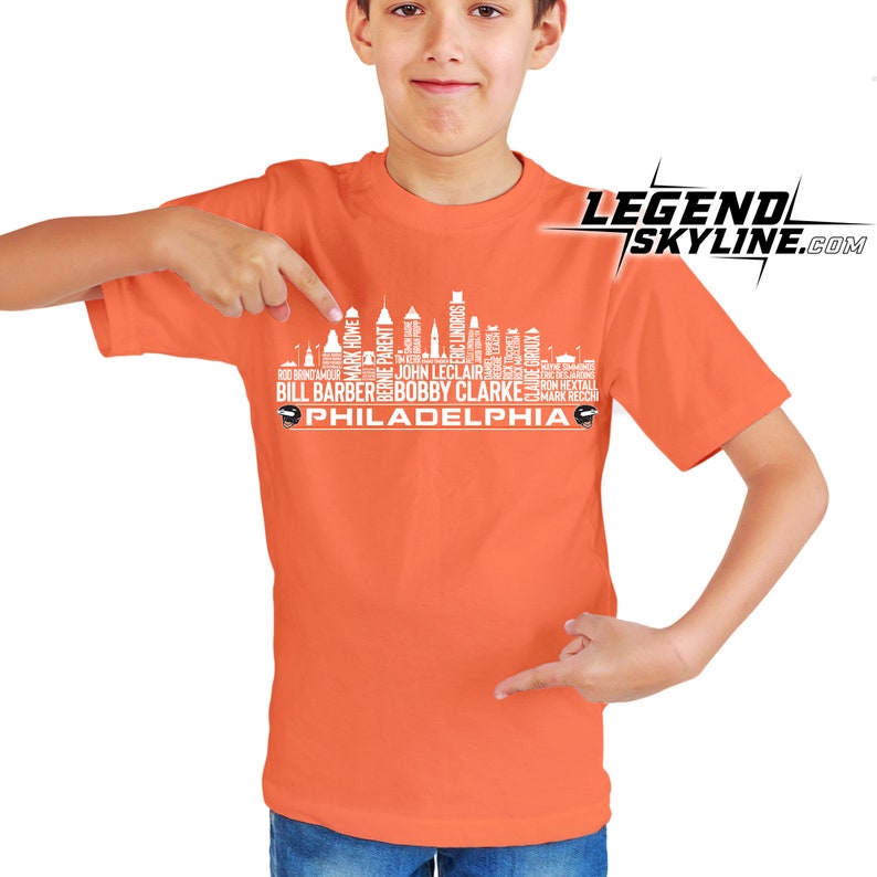 Może przedstawiać: Pomarańczowa koszulka z białym nadrukiem przedstawiającym panoramę Filadelfii, utworzoną z nazwisk znanych graczy Philadelphia Eagles. Pod panoramą znajduje się napis "PHILADELPHIA". Koszulkę nosi młoda osoba.
