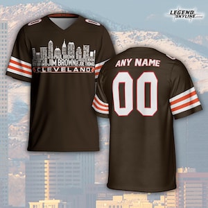 以下が含まれることがあります： 茶色のフットボールジャージ。袖には白とオレンジのストライプが入っています。前面には、クリーブランド・ブラウンズの伝説の名前と「JIM BROWN CLEVELAND」の文字が入ったスカイライングラフィックがあります。背面には数字「00」と「ANY NAME」の文字があります。
