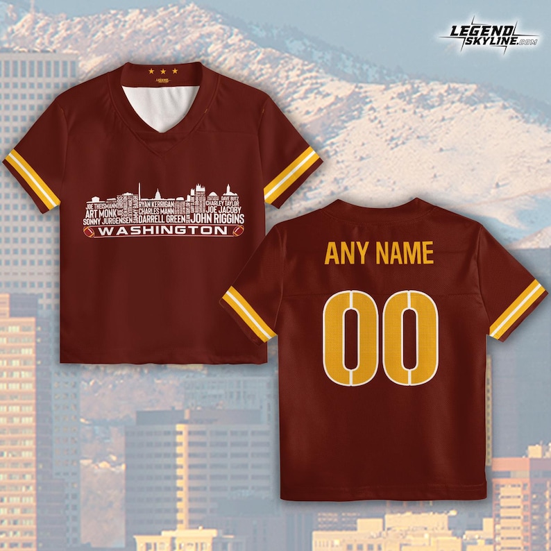 Peut inclure: Maillot de football marron avec des rayures dor&eacute;es sur les manches. Le devant pr&eacute;sente un graphique de skyline blanc avec des noms de joueurs et le texte "WASHINGTON". Le dos affiche "ANY NAME" et le num&eacute;ro "00" en or.