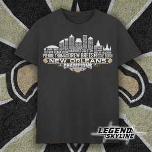 May include: Dark gray t-shirt featuring a white skyline design with player names and the text "New Orleans Champions." The shirt has a crew neck and short sleeves. The "Legend Skyline" logo is at the bottom.