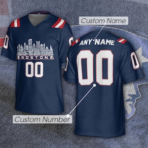 May include: A navy blue jersey with red and white shoulder stripes. The front features a white Boston skyline graphic with player names, the text BOSTON, and the number 00. The back displays the text ANY NAME and the number 00 in white.
