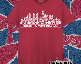 Może przedstawiać: Czerwona koszulka z białym nadrukiem przedstawiającym panoramę Filadelfii. Panorama składa się z nazwisk znanych zawodników baseballowych Philadelphia Phillies. Tekst "PHILADELPHIA" znajduje się na dole nadruku.