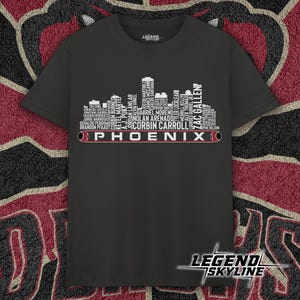 May include: A dark grey t-shirt with a white skyline design and player names, plus the word "PHOENIX" in white and red. The shirt has a crew neck and short sleeves. The "LEGEND SKYLINE" logo is at the bottom.