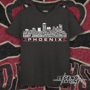 May include: A dark gray t-shirt with a white silhouette of the Phoenix skyline. The skyline is made up of the names of famous baseball players who have played for the Arizona Diamondbacks. The text "PHOENIX" is printed below the skyline.