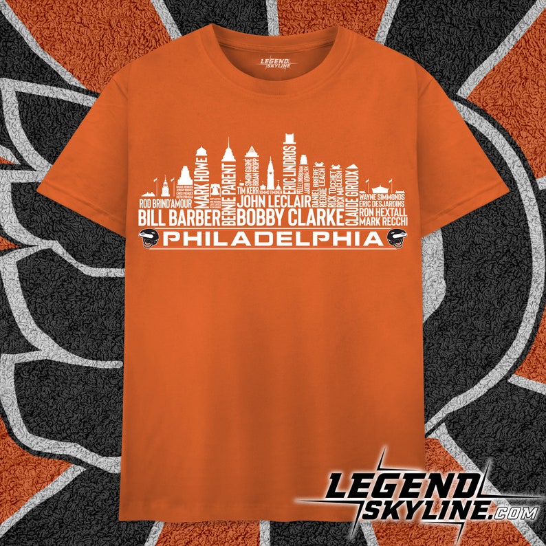Może przedstawiać: Pomarańczowy T-shirt z białym nadrukiem przedstawiającym panoramę Filadelfii. Nadruk składa się z nazwisk znanych hokeist&oacute;w Philadelphia Flyers. Na dole nadruku znajduje się napis "PHILADELPHIA".