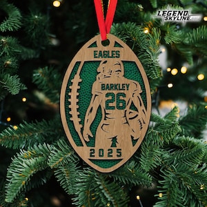 May include: Wooden football-shaped Christmas ornament with a red ribbon. The ornament features the text "EAGLES" and "BARKLEY 26 2025". The background is green, and the ornament is hanging on a Christmas tree.