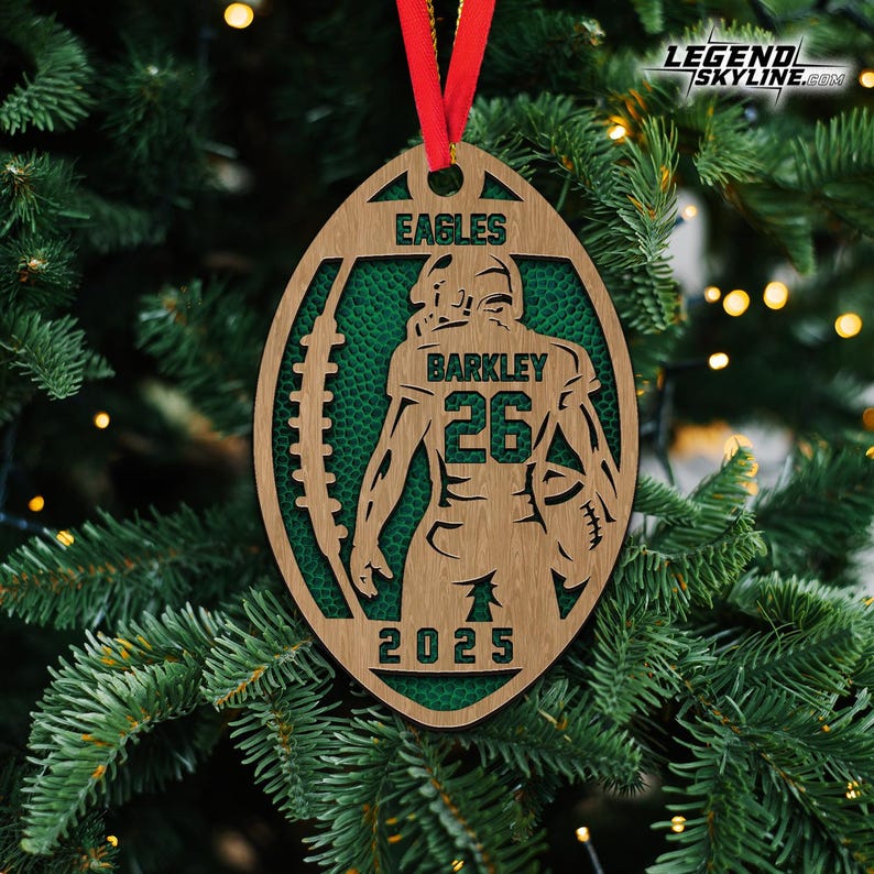 May include: Wooden football-shaped Christmas ornament with a red ribbon. The ornament features the text "EAGLES" and "BARKLEY 26 2025". The background is green, and the ornament is hanging on a Christmas tree.