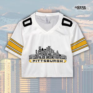 May include: White cropped football jersey with black and gold accents. The jersey features a V-neck and the word "PITTSBURGH" below a skyline graphic with player names. The sleeves have gold stripes.