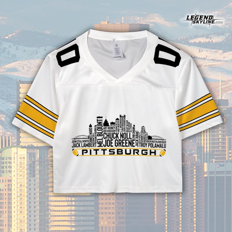 May include: White cropped football jersey with black and gold accents. The jersey features a V-neck and the word "PITTSBURGH" below a skyline graphic with player names. The sleeves have gold stripes.