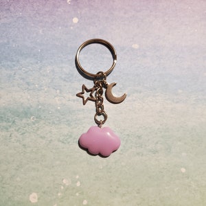 May include: A silver-toned keychain with a ring, a star, a crescent moon, and a lavender cloud charm. The star and moon charms are silver. The cloud charm is a soft, pastel purple color. The keychain is designed to hold keys or be used as a decorative accessory.