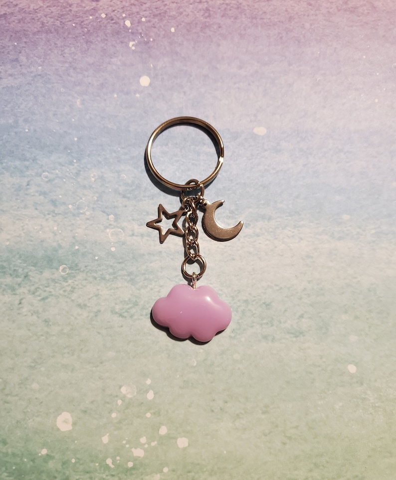 May include: A silver-toned keychain with a ring, a star, a crescent moon, and a lavender cloud charm. The star and moon charms are silver. The cloud charm is a soft, pastel purple color. The keychain is designed to hold keys or be used as a decorative accessory.