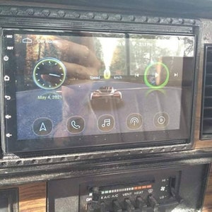 May include: A black car stereo with a touchscreen display showing a navigation map, a clock, and a speed reading. The screen also shows icons for phone, music, and a microphone. The stereo is installed in a car dashboard with woodgrain trim.