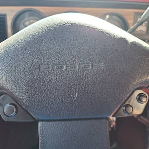 May include: A black leather steering wheel with the word "DODGE" embossed in the center. The steering wheel has two black buttons on each side.
