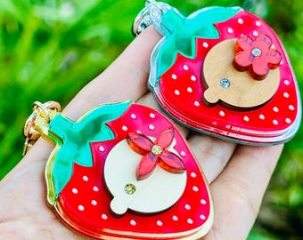 May include: Two strawberry-shaped keychains with red enamel, white dots, and green tops. Each keychain features a wooden circle with a red flower and a small gem. The keychains have gold-colored metal trim.