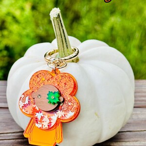 May include: A tan and cherry orange glitter pumpkin charm with a gold key ring attached to a white pumpkin. The charm has a brown face with a green leaf on top.