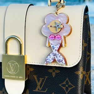 May include: A white and brown designer handbag with a gold lock and a mermaid charm. The charm is pink and white with glitter and a starfish. The bag is on a sandy beach.