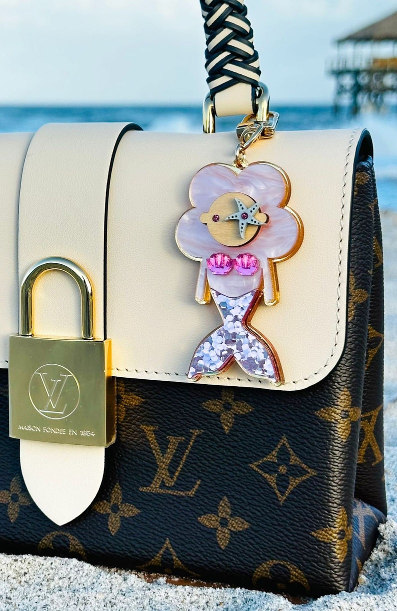 May include: A white and brown designer handbag with a gold lock and a mermaid charm. The charm is pink and white with glitter and a starfish. The bag is on a sandy beach.