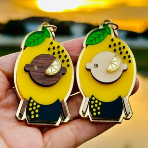 May include: Two yellow lemon-shaped keychains with black and gold accents. One keychain has a brown face and the other has a white face. Both keychains have a green leaf on top.