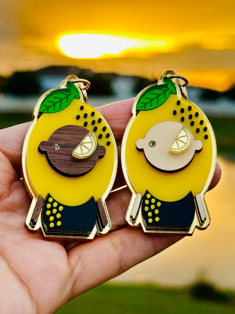 May include: Two yellow lemon-shaped keychains with black and gold accents. One keychain has a brown face and the other has a white face. Both keychains have a green leaf on top.