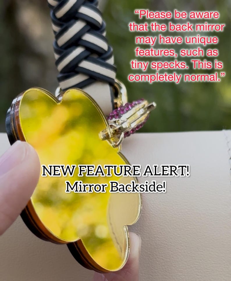 May include: A gold-colored heart-shaped mirror with a black border. The mirror is attached to a black and white braided strap. The text "NEW FEATURE ALERT! Mirror Backside!" is printed on the image. The text "Please be aware that the back mirror may have unique features, such as tiny specks. This is completely normal." is also printed on the image.