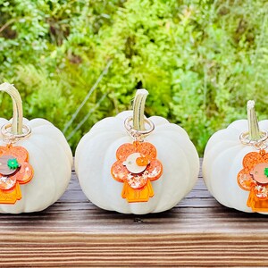 May include: Three white pumpkins with orange and green glitter flower charms attached to them. The charms are shaped like a person with a flower head.