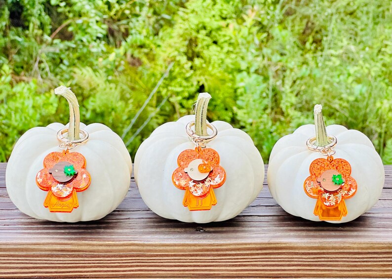 May include: Three white pumpkins with orange and green glitter flower charms attached to them. The charms are shaped like a person with a flower head.