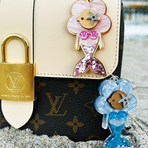 May include: Two mermaid-themed keychains with glitter accents. The keychain on the left is pink with a silver starfish, and the keychain on the right is blue with a blue starfish. Both keychains are attached to a brown and white patterned handbag.
