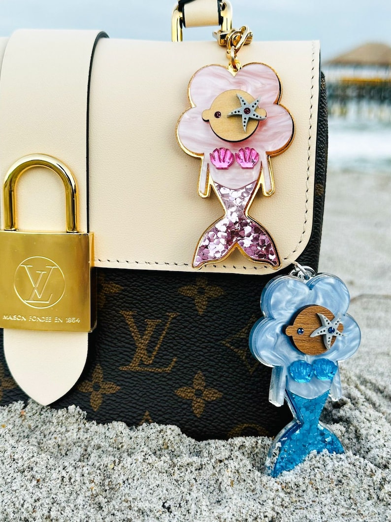 May include: Two mermaid-themed keychains with glitter accents. The keychain on the left is pink with a silver starfish, and the keychain on the right is blue with a blue starfish. Both keychains are attached to a brown and white patterned handbag.