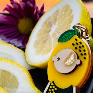 May include: A yellow and black lemon-shaped resin brooch with a gold-tone metal pin. The brooch features a small, white, resin lemon slice in the center. The brooch is sitting on top of a lemon slice.
