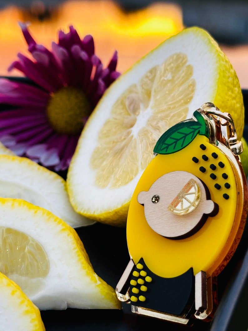 May include: A yellow and black lemon-shaped resin brooch with a gold-tone metal pin. The brooch features a small, white, resin lemon slice in the center. The brooch is sitting on top of a lemon slice.