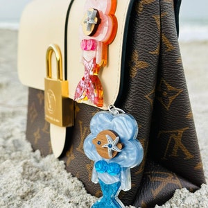 May include: Two mermaid-themed keychains with a pink and blue iridescent finish. The pink keychain has a starfish and a pink seashell. The blue keychain has a starfish and a blue seashell. Both keychains are attached to a brown and white handbag.