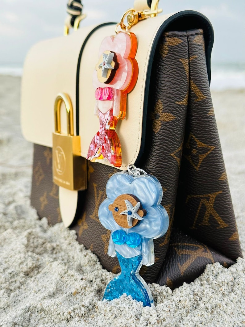 May include: Two mermaid-themed keychains with a pink and blue iridescent finish. The pink keychain has a starfish and a pink seashell. The blue keychain has a starfish and a blue seashell. Both keychains are attached to a brown and white handbag.