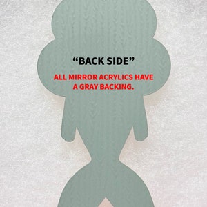 May include: Back side of a light blue mermaid-shaped acrylic ornament. The text "BACK SIDE" and "ALL MIRROR ACRYLICS HAVE A GRAY BACKING." is printed in red.