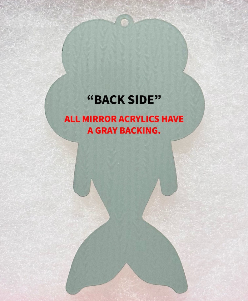 May include: Back side of a light blue mermaid-shaped acrylic ornament. The text "BACK SIDE" and "ALL MIRROR ACRYLICS HAVE A GRAY BACKING." is printed in red.