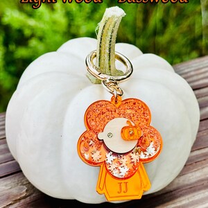May include: Orange and white glitter pumpkin charm with a wood face and the initials "JJ" on a gold key chain. The charm is hanging from a white pumpkin.