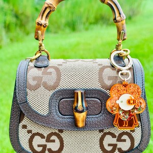 May include: A brown and beige Gucci handbag with a bamboo handle and a brown leather trim. The bag has a GG pattern and a bamboo closure. A small orange flower charm with the initials "JJ" is attached to the bag.