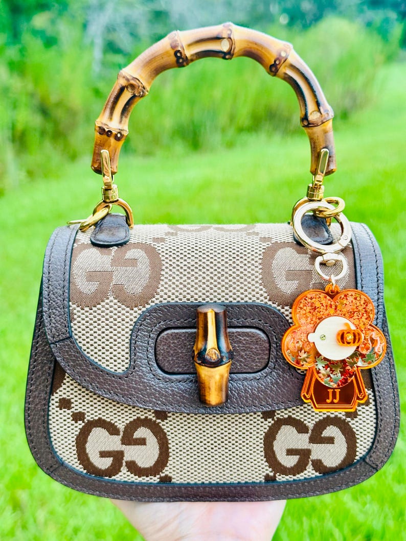 May include: A brown and beige Gucci handbag with a bamboo handle and a brown leather trim. The bag has a GG pattern and a bamboo closure. A small orange flower charm with the initials "JJ" is attached to the bag.