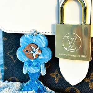 May include: A blue mermaid charm with a starfish and a blue eye. The charm is attached to a white and brown handbag with a gold lock that says "Maison Fond&eacute;e en 1854".