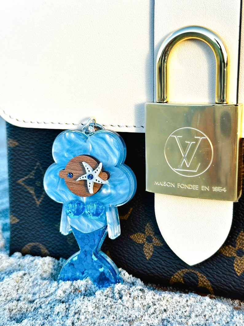 May include: A blue mermaid charm with a starfish and a blue eye. The charm is attached to a white and brown handbag with a gold lock that says "Maison Fond&eacute;e en 1854".