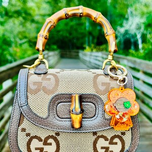 May include: A brown and beige Gucci handbag with a bamboo handle and a brown leather trim. The bag has a GG pattern and a gold-toned buckle. A small orange and green flower charm is attached to the bag with the word "Angie" written on it.