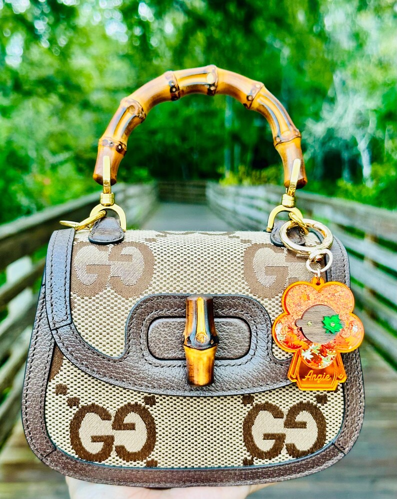 May include: A brown and beige Gucci handbag with a bamboo handle and a brown leather trim. The bag has a GG pattern and a gold-toned buckle. A small orange and green flower charm is attached to the bag with the word "Angie" written on it.