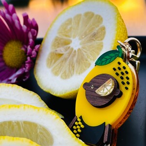 May include: A yellow lemon-shaped keychain with a black and brown design. The keychain features a small lemon slice and black polka dots. The keychain is surrounded by lemon slices.
