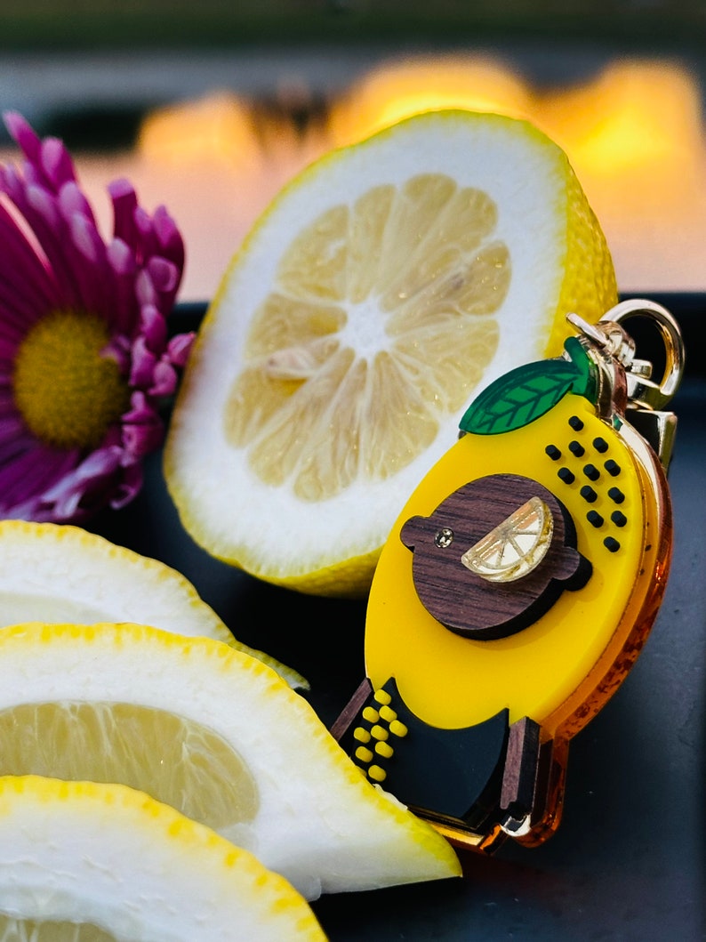 May include: A yellow lemon-shaped keychain with a black and brown design. The keychain features a small lemon slice and black polka dots. The keychain is surrounded by lemon slices.